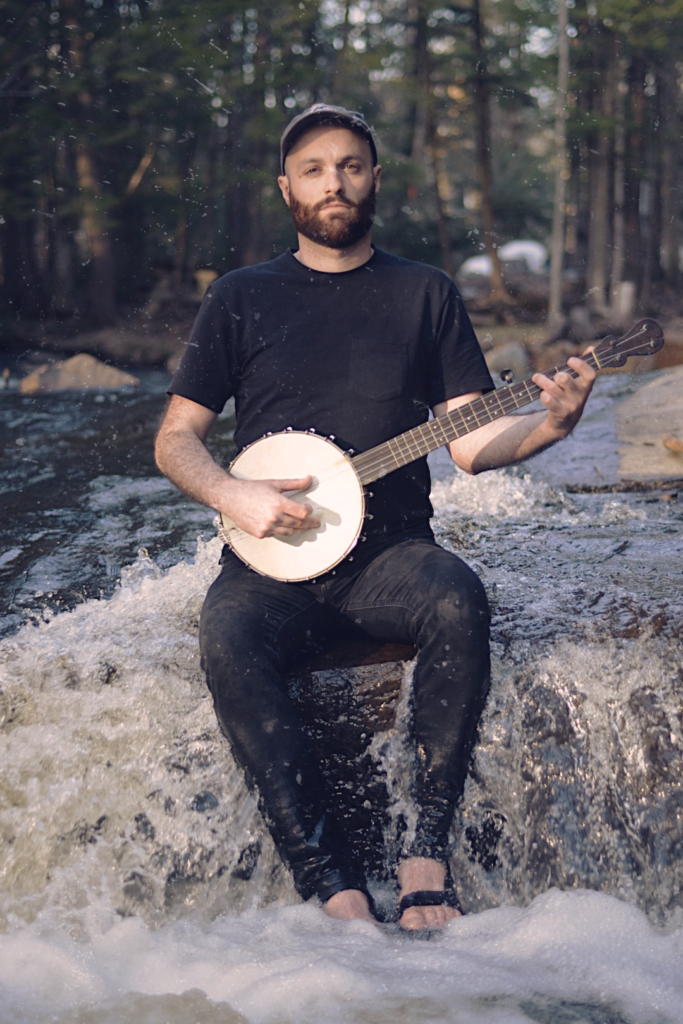 Ben Nesrallah (Hallarsen) sits on a rock in a small rushing river playing a 5-string banjo.